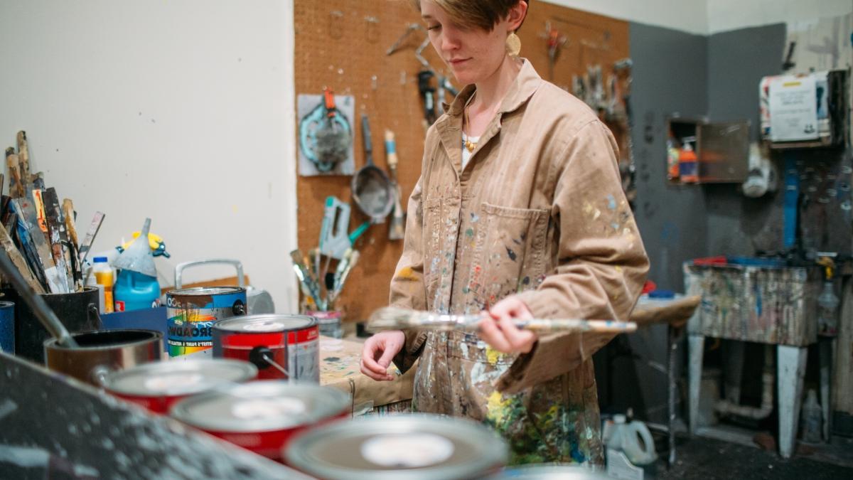 Student holds a paintbrush in front of a row of paint cans.